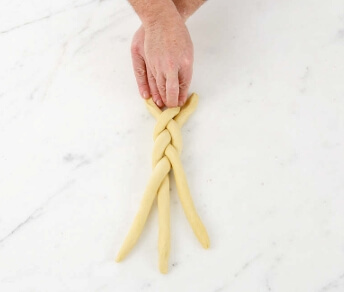 Three floured strands of brioche dough being braided together on a clean work surface, ends tucked neatly underneath.
