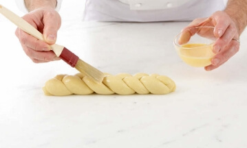 Braided brioche loaf brushed with egg wash, sprinkled with pearl sugar, resting on a tray before baking.
