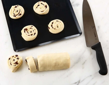 Slices of rolled dough, filled with pastry cream and raisins, being cut into individual portions to create perfectly shaped raisin buns.
