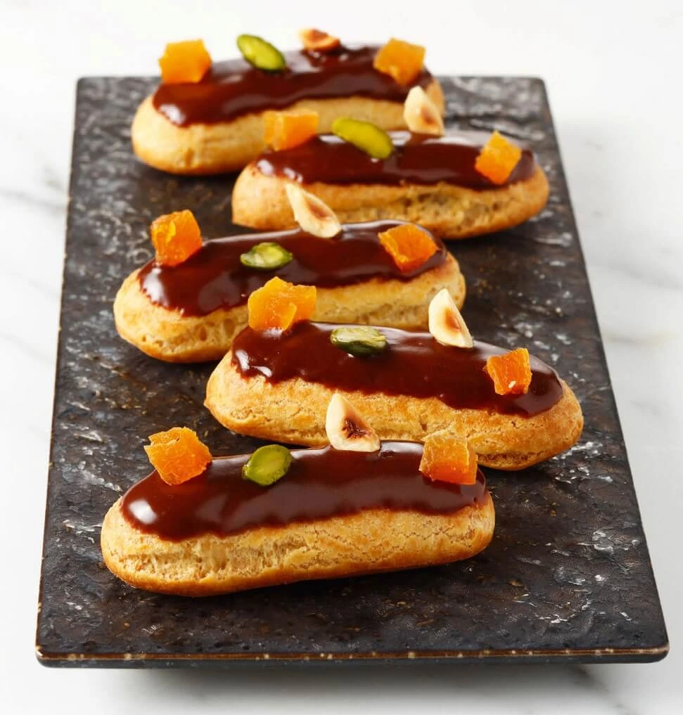 A close-up of mini éclairs topped with chocolate fondant, pistachios, dried apricots, and toasted hazelnuts, showcasing their glossy finish.