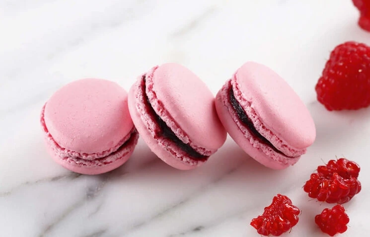 Raspberry macarons with smooth pink shells and vibrant raspberry confit filling, stacked on a white plate.