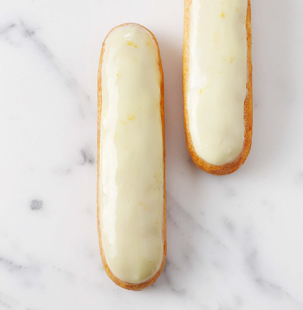 Close-up of a lemon éclair topped with crunchy craquelin, filled with smooth lemon cream, on a white plate.