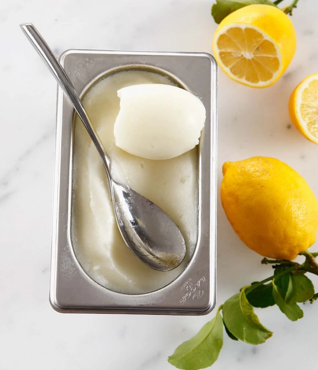 Close-up of a creamy, bright yellow lemon sorbet scooped into a glass bowl, garnished with fresh lemon zest and mint leaves, showcasing its smooth texture and vibrant color.
