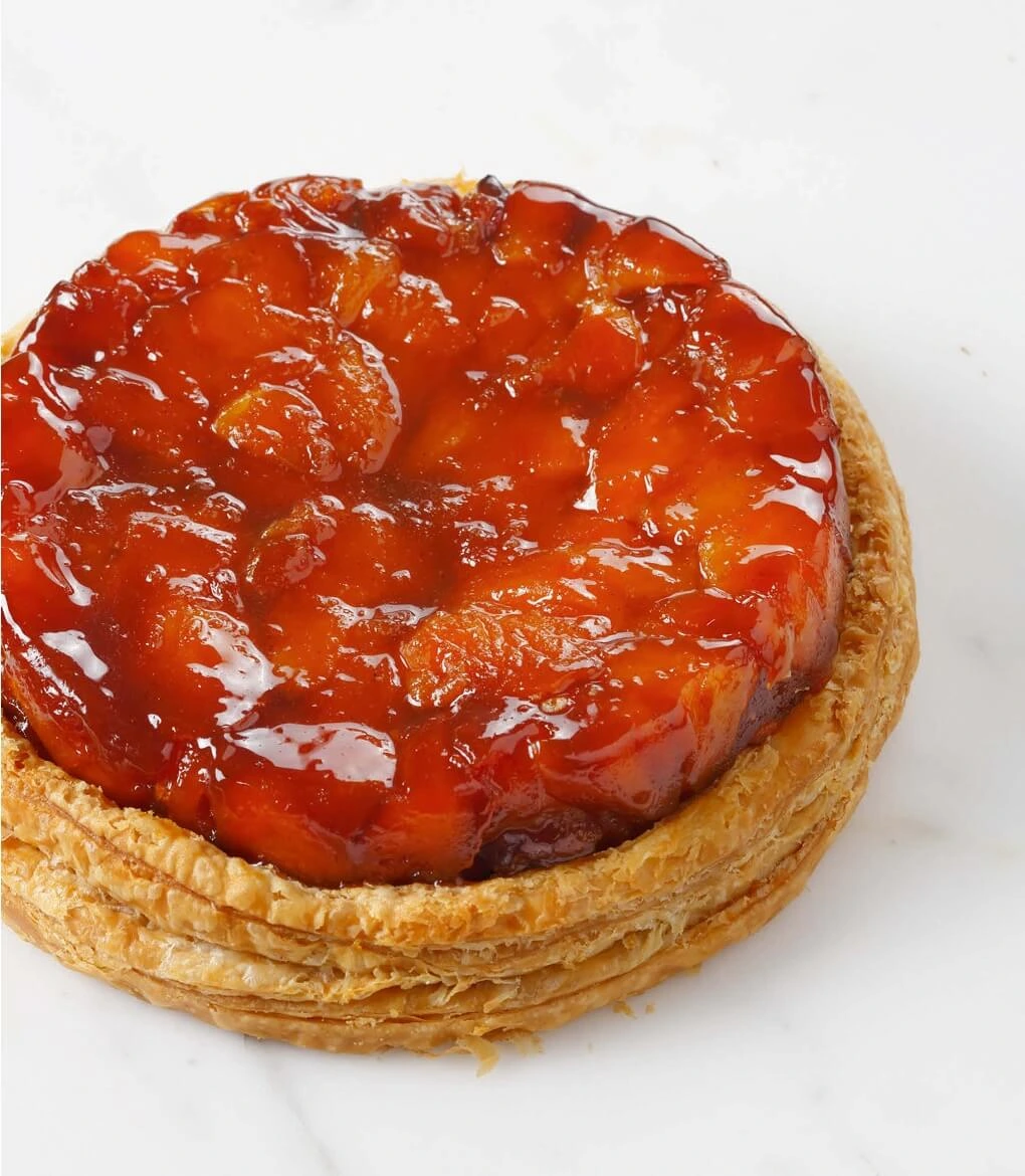 Close-up of a golden caramelized Tarte Tatin topped with glossy apple slices on a flaky puff pastry base, served on a rustic wooden board.