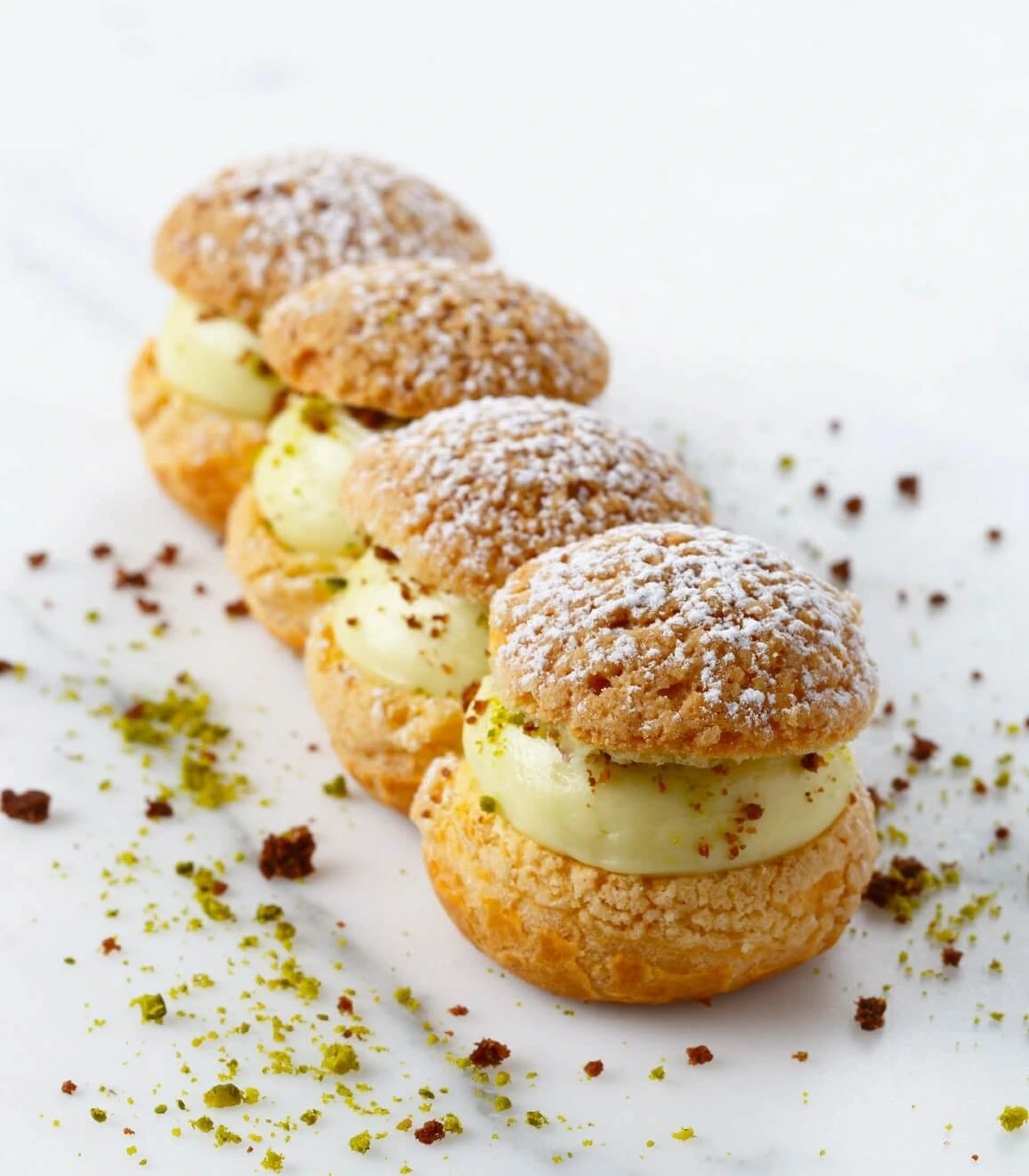 Close-up of a Pistachoux pastry showing a golden craquelin topping, creamy pistachio filling, and a vibrant lemon cream insert in the center.