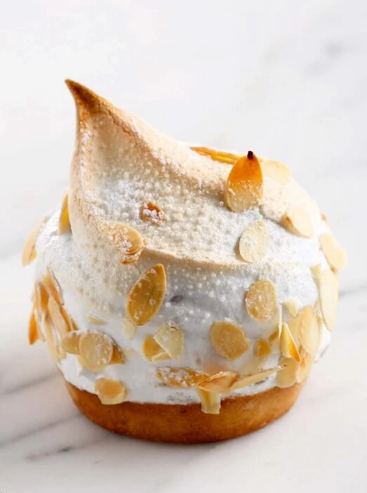 Close-up of a golden brioche tartlet topped with glossy Italian meringue and toasted almond slices, with visible layers of syrup-soaked brioche and creamy pastry filling.