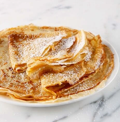 Freshly cooked crêpes folded on a serving plate, topped with powdered sugar and garnished with berries, steaming gently.