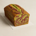 A high-resolution photo of a marbled pistachio and raspberry loaf cake, sliced to reveal vibrant green and pink swirls inside a golden-brown crust. The cake sits on a parchment-lined wooden board, garnished with crushed pistachios and fresh raspberries.